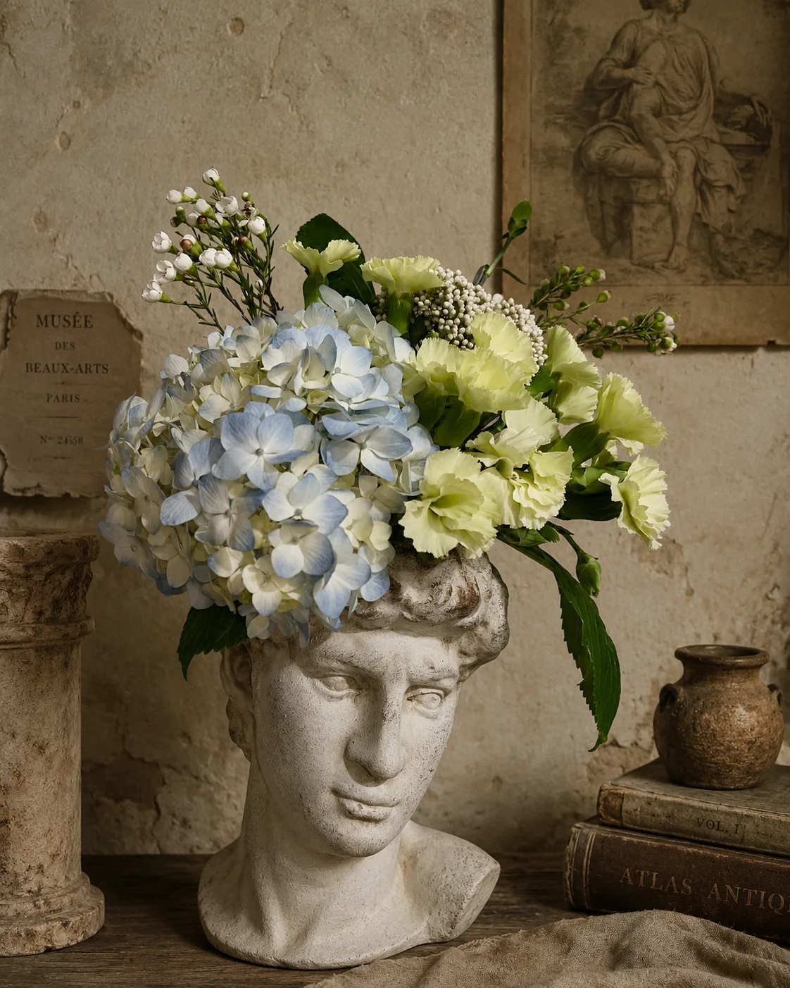 Blue hydrangea and pale yellow carnation arrangement in a classical bust-shaped vase, styled with antique books and a vintage museum postcard
