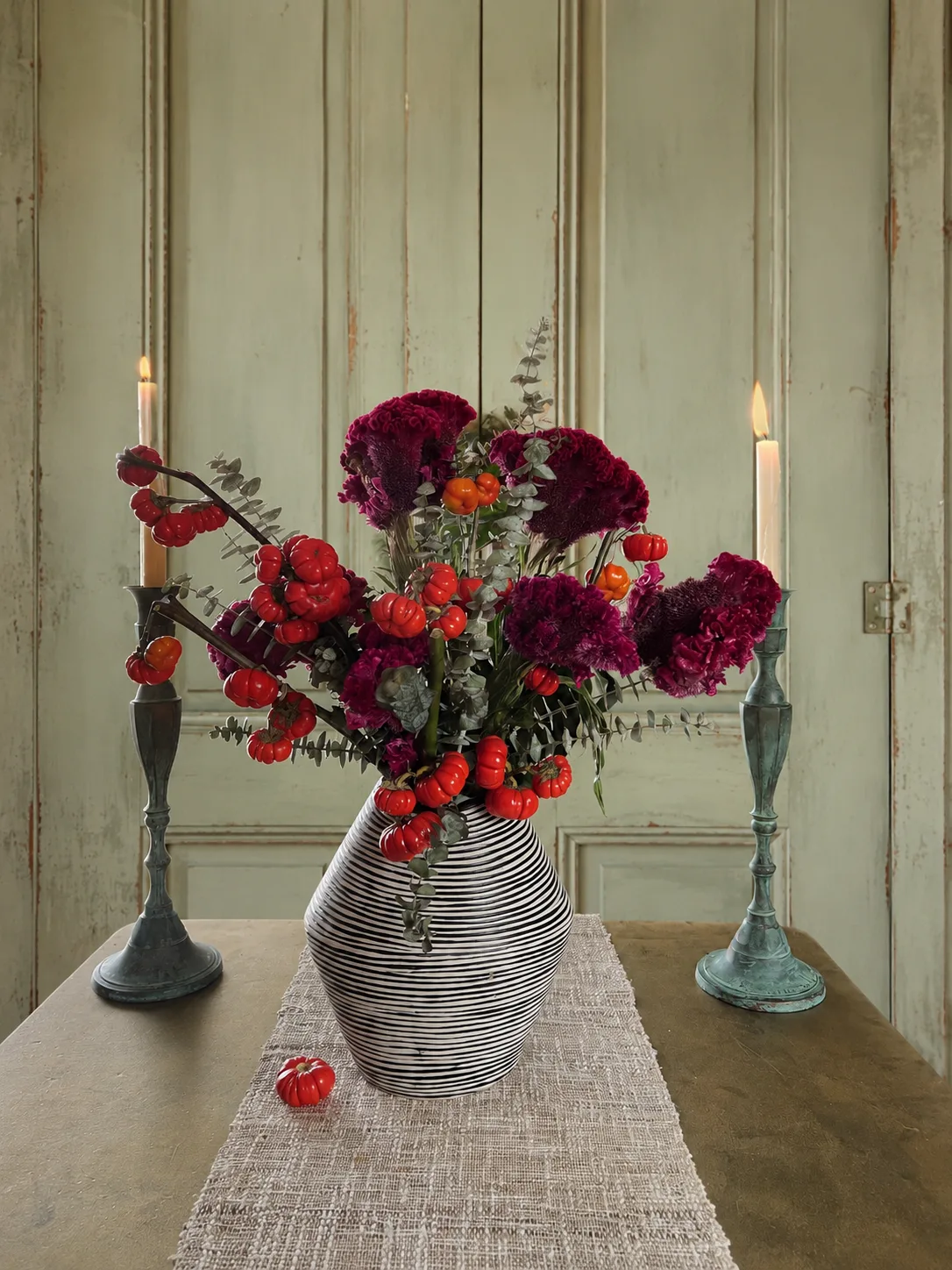 Magenta celosia and red berry arrangement in a striped vase flanked by candlesticks