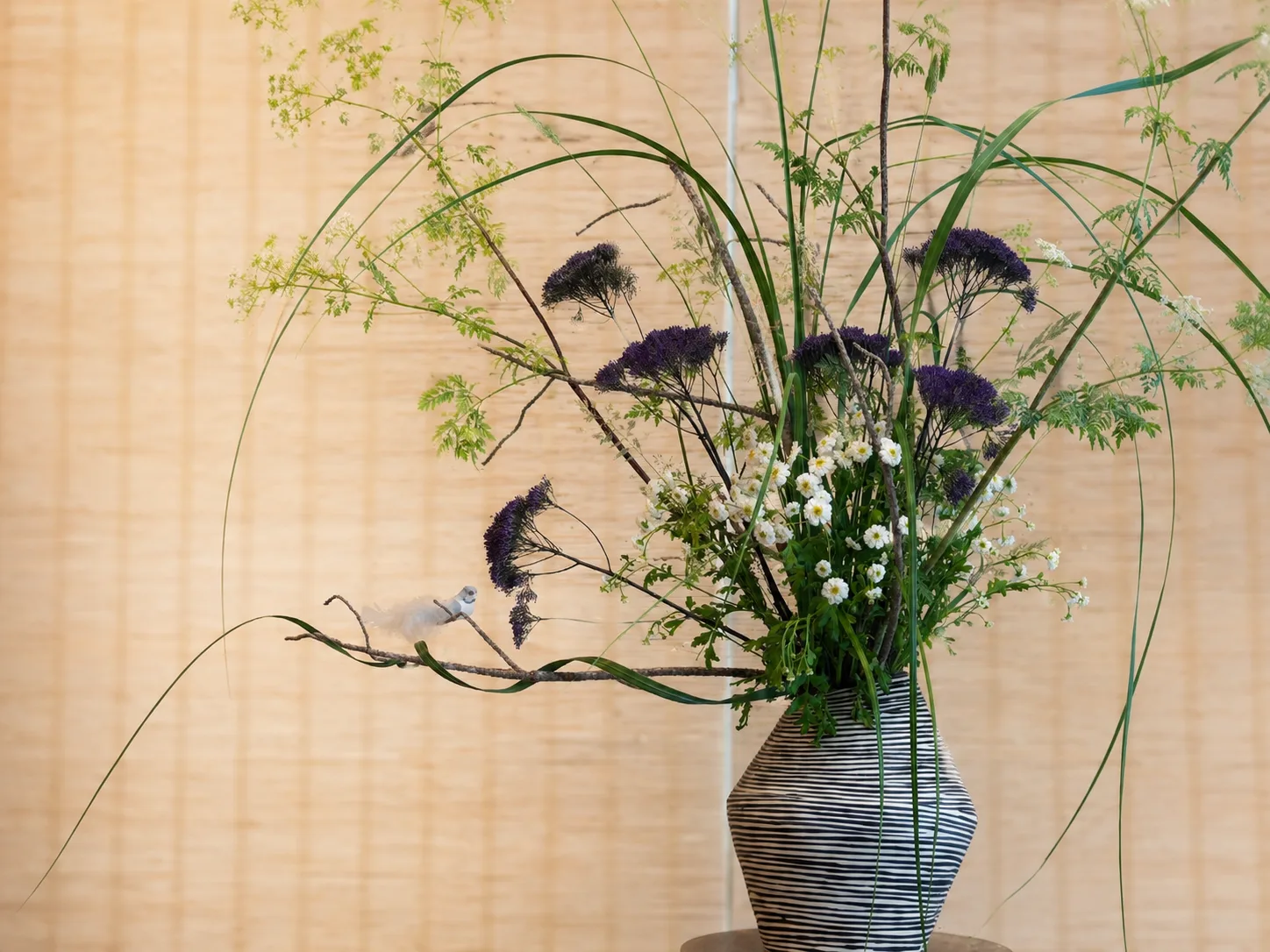 Wild grass and purple statice ikebana arrangement in a striped black-and-white vase against a woven wall screen