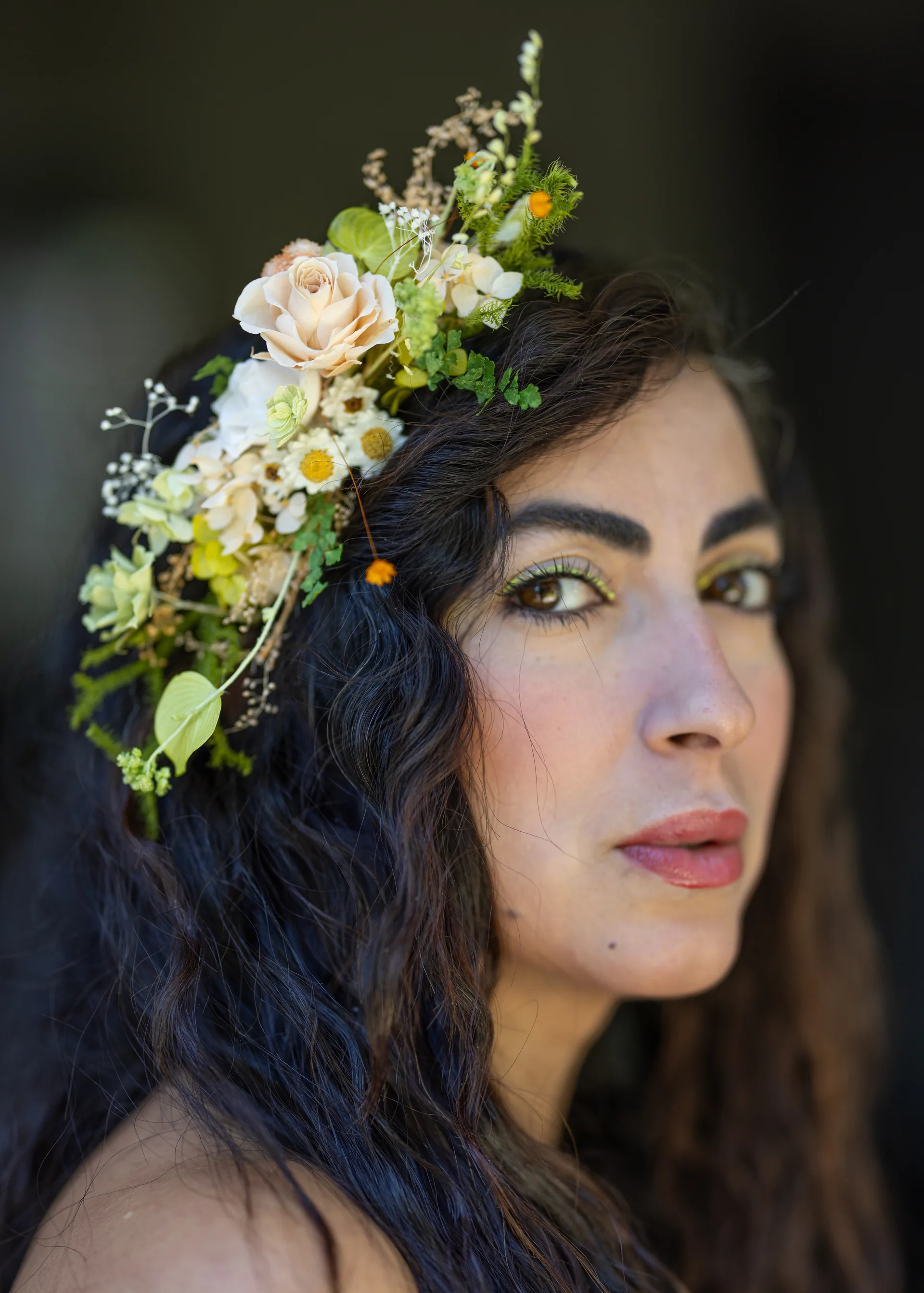 Woman in profile with a large asymmetric flower crown of peach roses, cream hydrangea, and wild greenery cascading through her dark curled hair