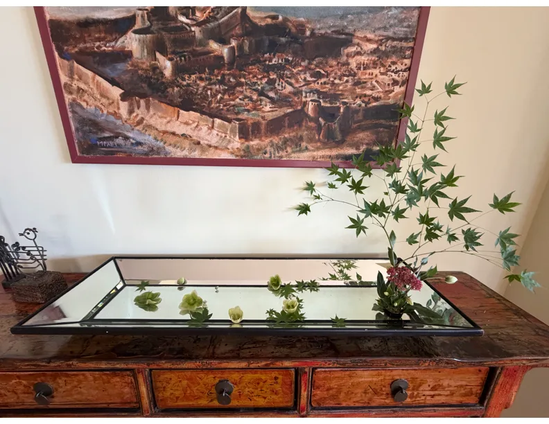 Japanese maple branch and small white blossoms arranged in a mirrored tray on an antique console table beneath a framed painting