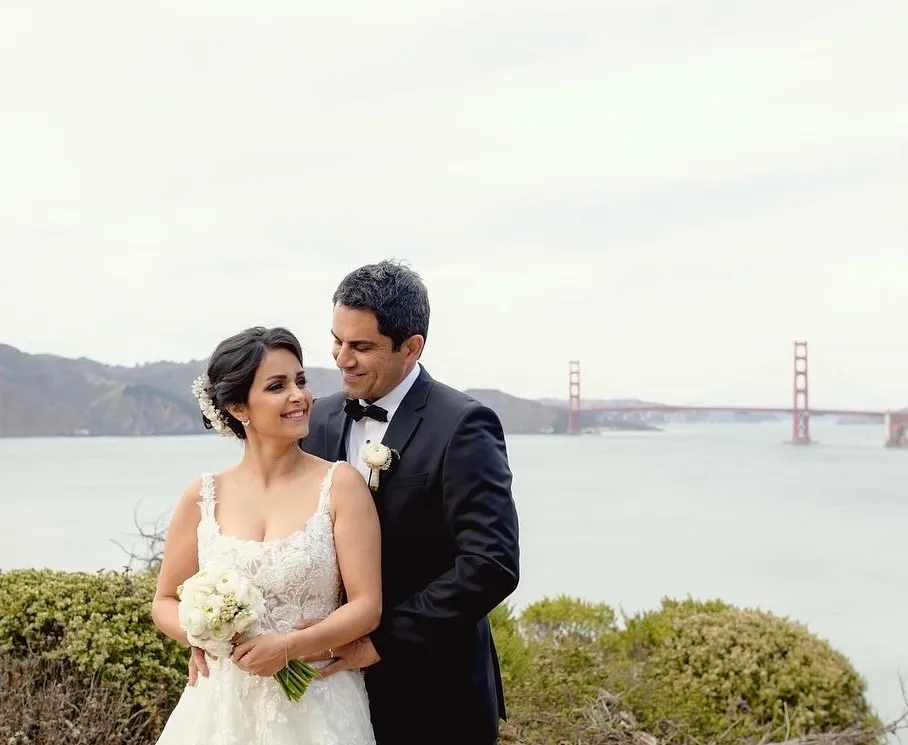 Bride and groom smiling at each other on a bluff overlooking the Golden Gate Bridge, the bride holding a white ranunculus and baby's breath bouquet