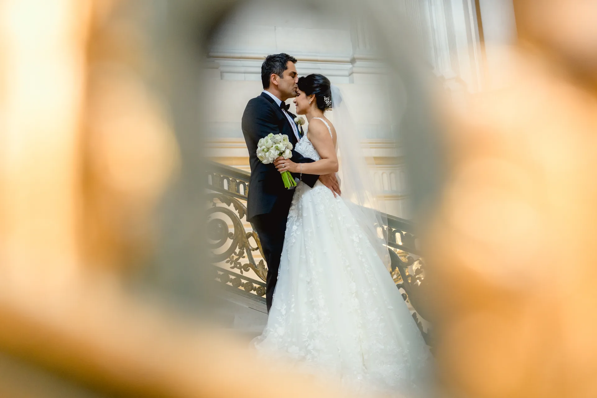 Bride and groom sharing a kiss on a grand staircase, seen framed through a blurred gold wedding ring